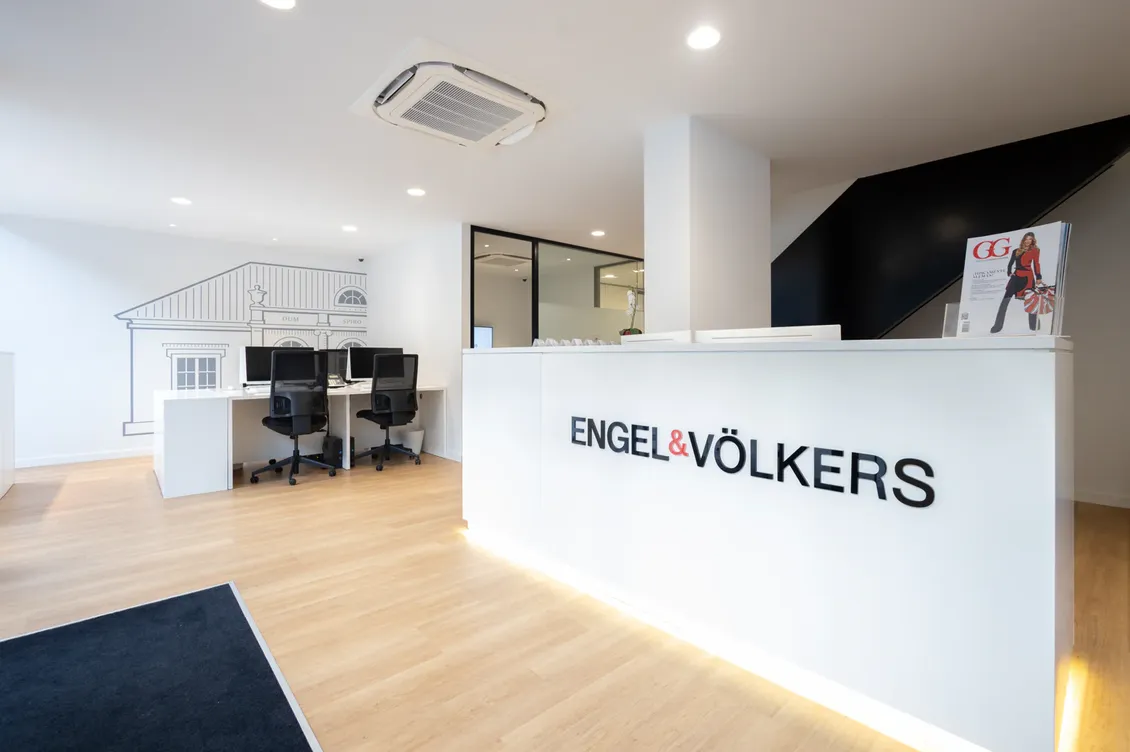 Recepción de las oficinas de Engel & Völkers con escritorio blanco, logotipo negro y diseño moderno. Al fondo se ven escritorios con ordenadores.