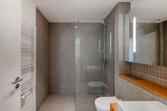 Minimal modern bathroom with glass walk-in shower, grey square tiles, wall-mounted rain and handheld shower, white toilet, wooden shelf and mirrored cabinet.
