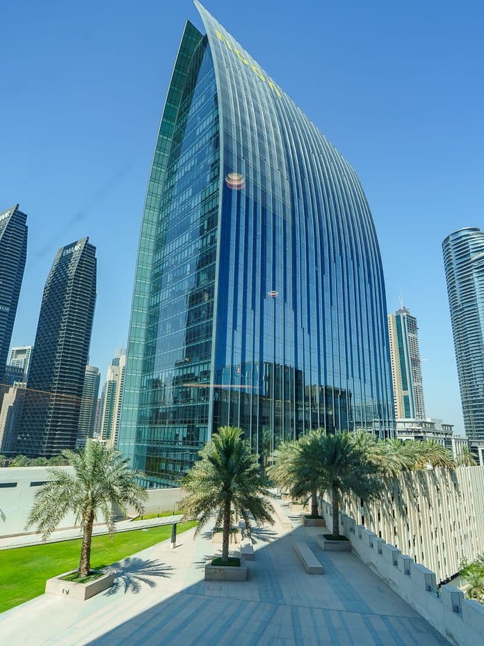 Exterior view of Boulevard Plaza tower surrounded by Dubai skyscrapers, seen from a landscaped terrace with palm trees and open walkways.