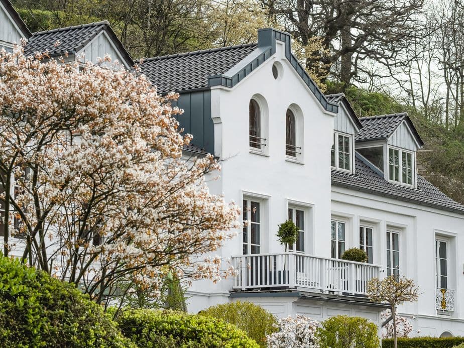 White house with a dark roof and dormer windows, surrounded by blooming trees and shrubs, set against a backdrop of bare trees.