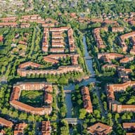 Luftaufnahme von Bergerdorf mit roten Dächern, baumbestandenen Straßen und Kanälen, umgeben von Grünflächen und anderen Häusern.