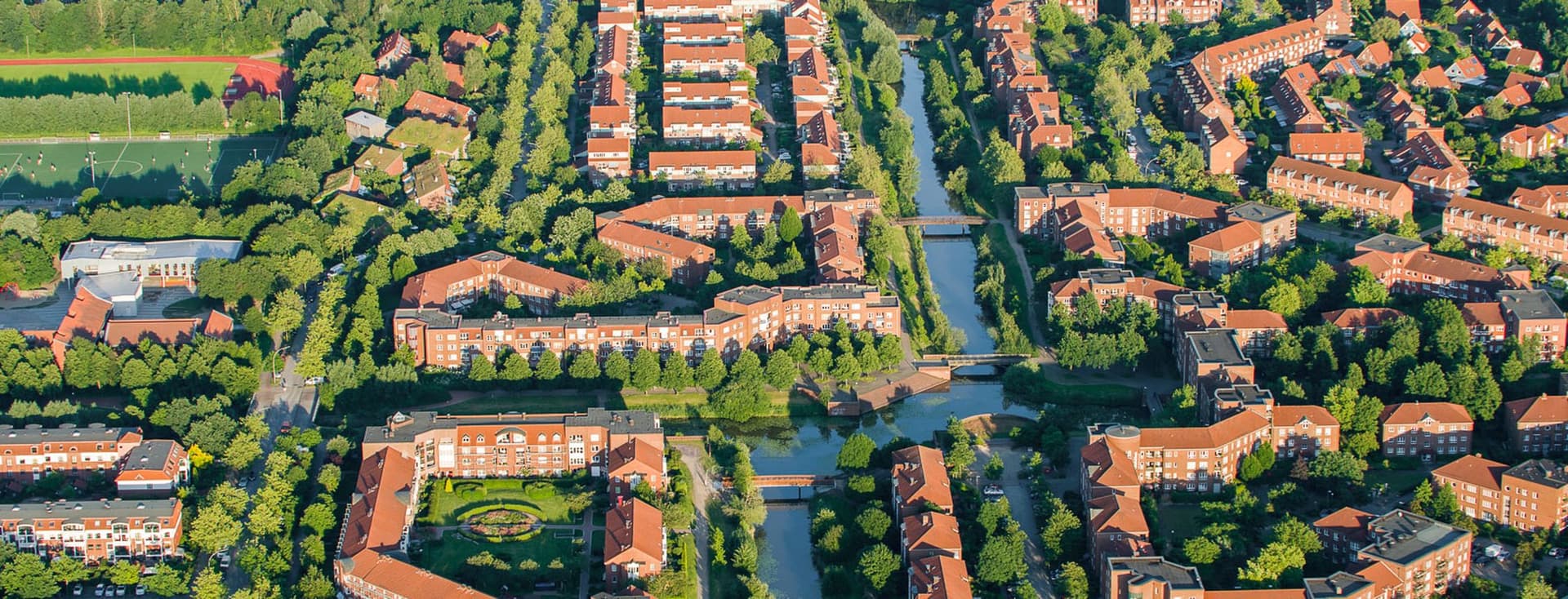 Aerial view of Bergerdorf with red roofs, tree-lined streets, and canals, surrounded by green spaces and other houses.