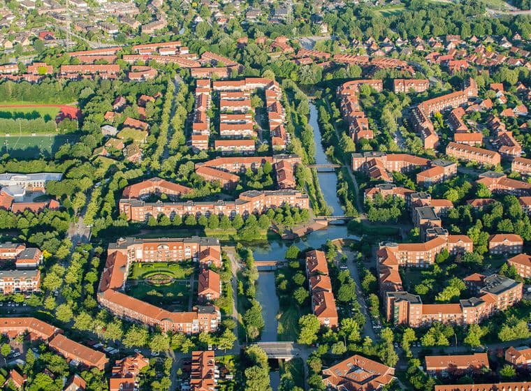 Aerial view of Bergerdorf with red roofs, tree-lined streets, and canals, surrounded by green spaces and other houses.
