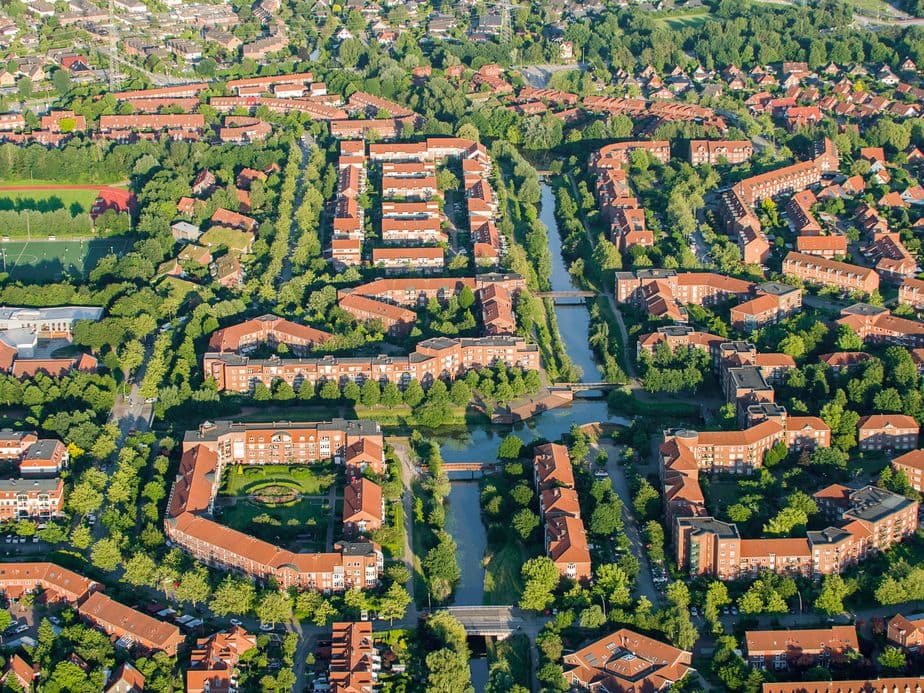 Aerial view of Bergerdorf with red-roofed buildings, tree-lined streets, and canals, surrounded by greenery and other houses.