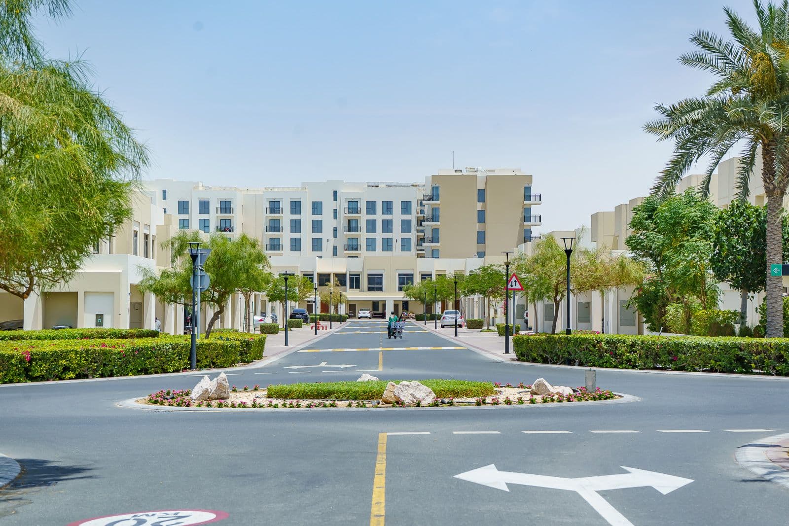 Hauptzufahrtsstraße in Town Square Dubai, die zu mittelgroßen Apartmentgebäuden führt, gesäumt von gepflegten Hecken, Palmen und moderner Straßenbeleuchtung.