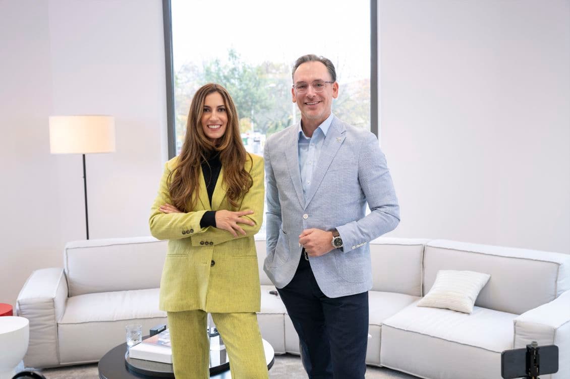 A young woman in a green two-piece suit with long brown hair and folded arms stands next to a middle-aged man in a light blue jacket, with a gray couch in the background.