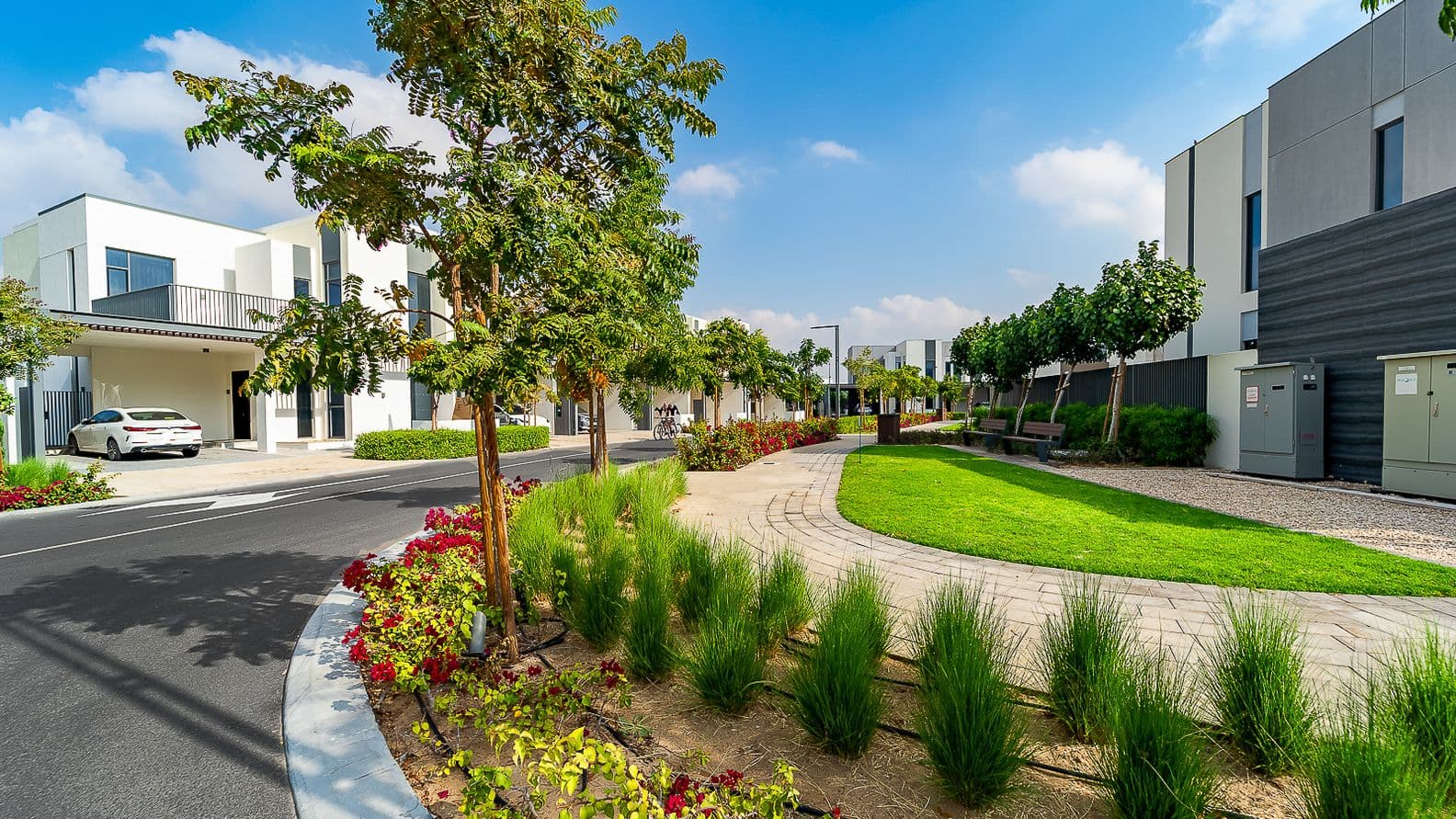 Blick auf Bodenhöhe auf einen angelegten Weg in Arabian Ranches 3, gesäumt von jungen Bäumen, Blumenbeeten, modernen Villen und Bänken unter einem klaren blauen Himmel.