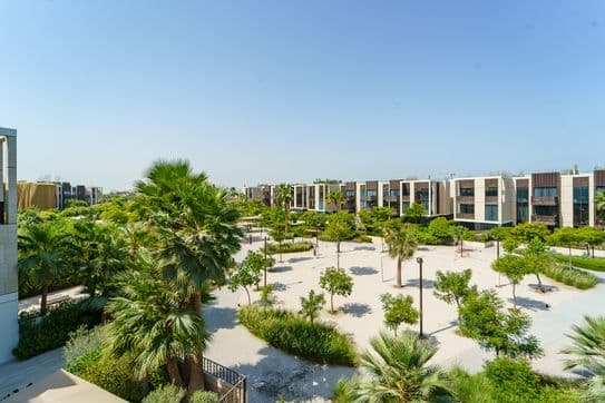 Moderne Wohngegend mit grünen Bäumen, Palmen und einem sandigen Weg unter klarem blauem Himmel. Zeitgenössische Gebäude bilden den Hintergrund.