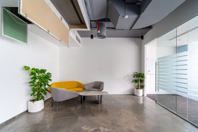 Elegant office lounge with light grey sofas, wooden partitions, glass panels, and soft lighting creating a comfortable, professional space for informal meetings.