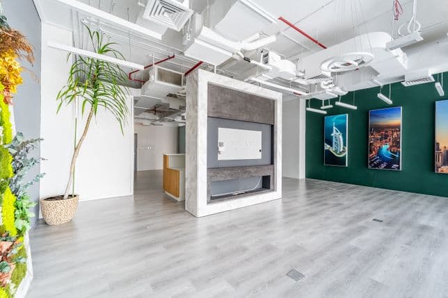 Bright modern showroom with central concrete display/TV niche, light wood floor, exposed ceiling, tall potted plant and green wall with city photos.