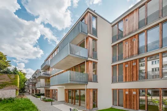 Modernes, mehrstöckiges Wohnhaus mit Holzverkleidung, großen Glasfenstern und Metallbalkonen, das an einen gepflasterten Gehweg mit grünem Rasen grenzt.