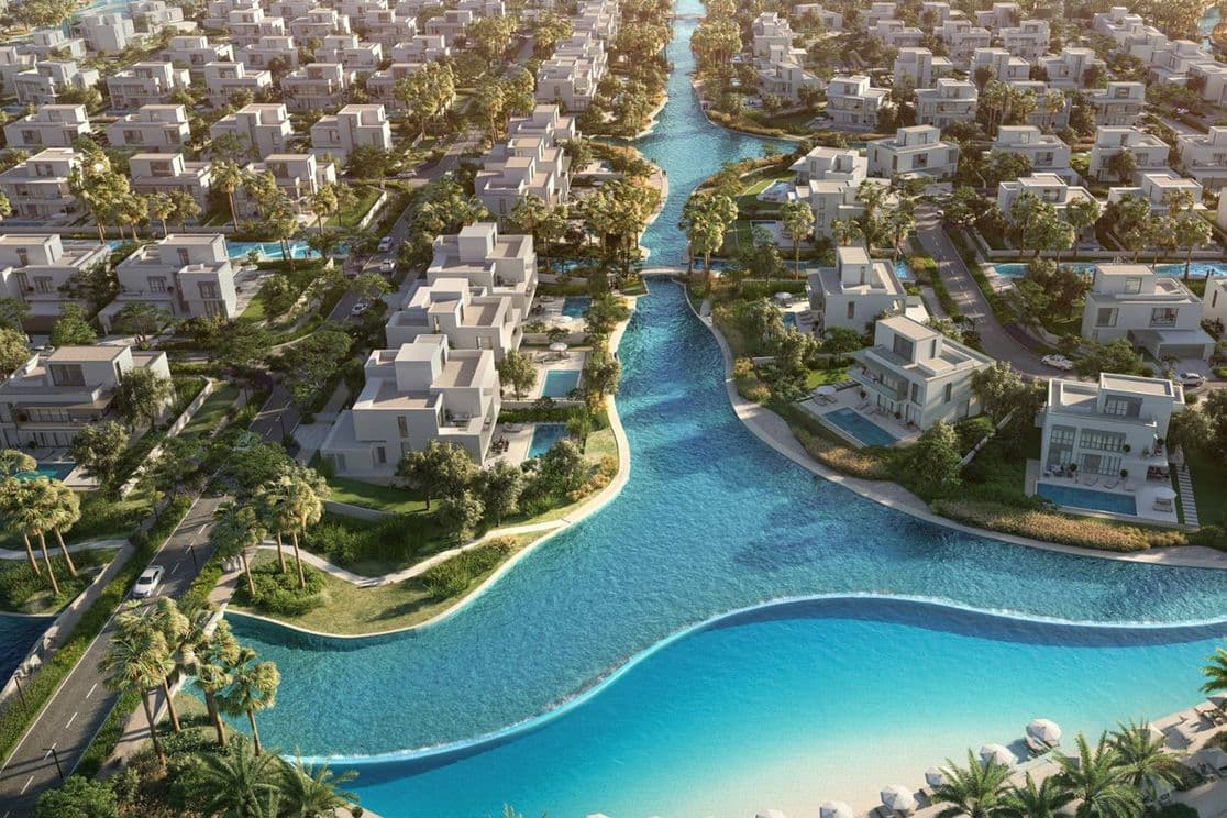 Luftaufnahme einer Wohnanlage mit modernen weißen Häusern, blauen Wasserwegen, Palmen und einem Sandstrand.