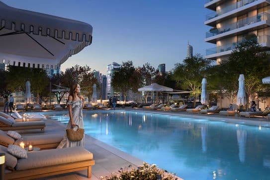 Woman in a dress stands by a lit pool at dusk, surrounded by lounge chairs and umbrellas, with city skyscrapers in the background.
