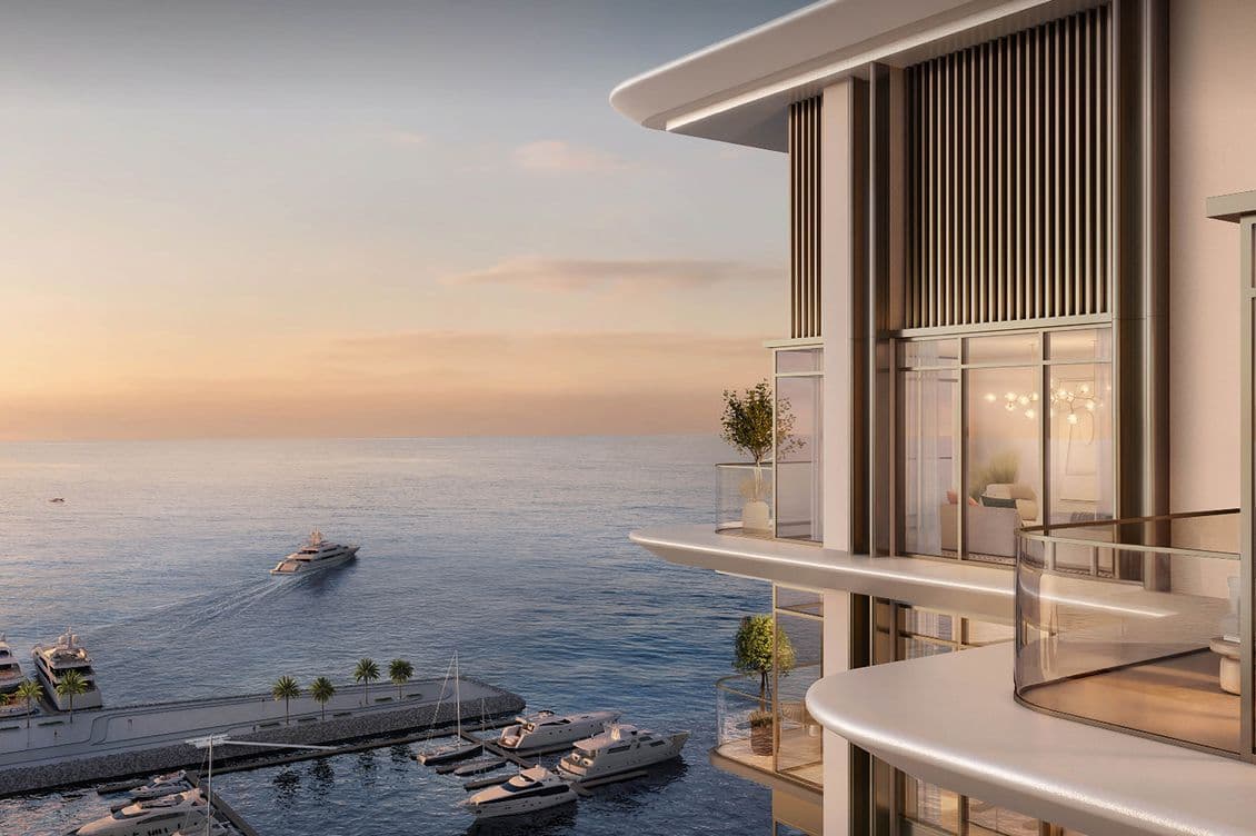 Modern waterfront building with balconies overlooking a marina and ocean at sunset. Boats are docked, and a yacht sails in the distance.