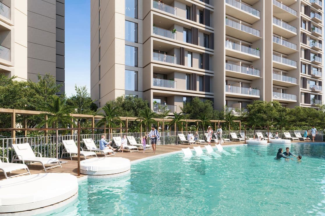 Outdoor swimming pool with lounge chairs and people relaxing. Tall apartment buildings and greenery in the background under a clear blue sky.