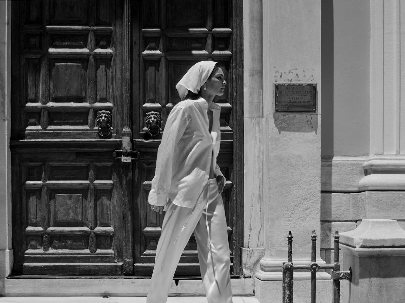 Μια ασπρόμαυρη φωτογραφία μιας γυναίκας με λευκά ρούχα που περπατά δίπλα από ένα κτίριο με μια μεγάλη ξύλινη πόρτα.
