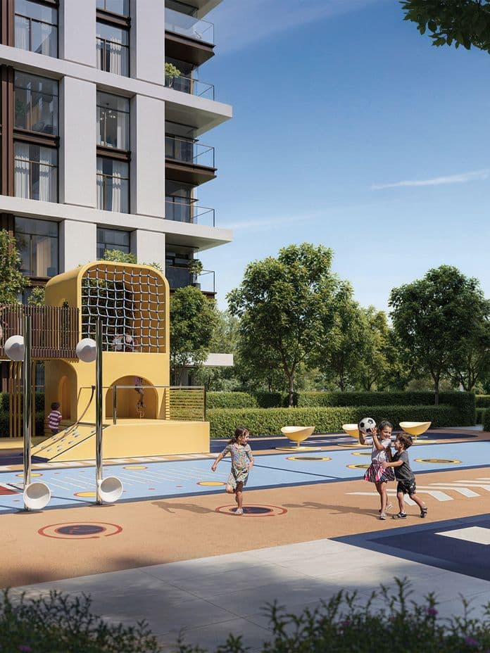 Children playing on a sunny day in a colorful playground near a modern apartment building, surrounded by greenery and trees.