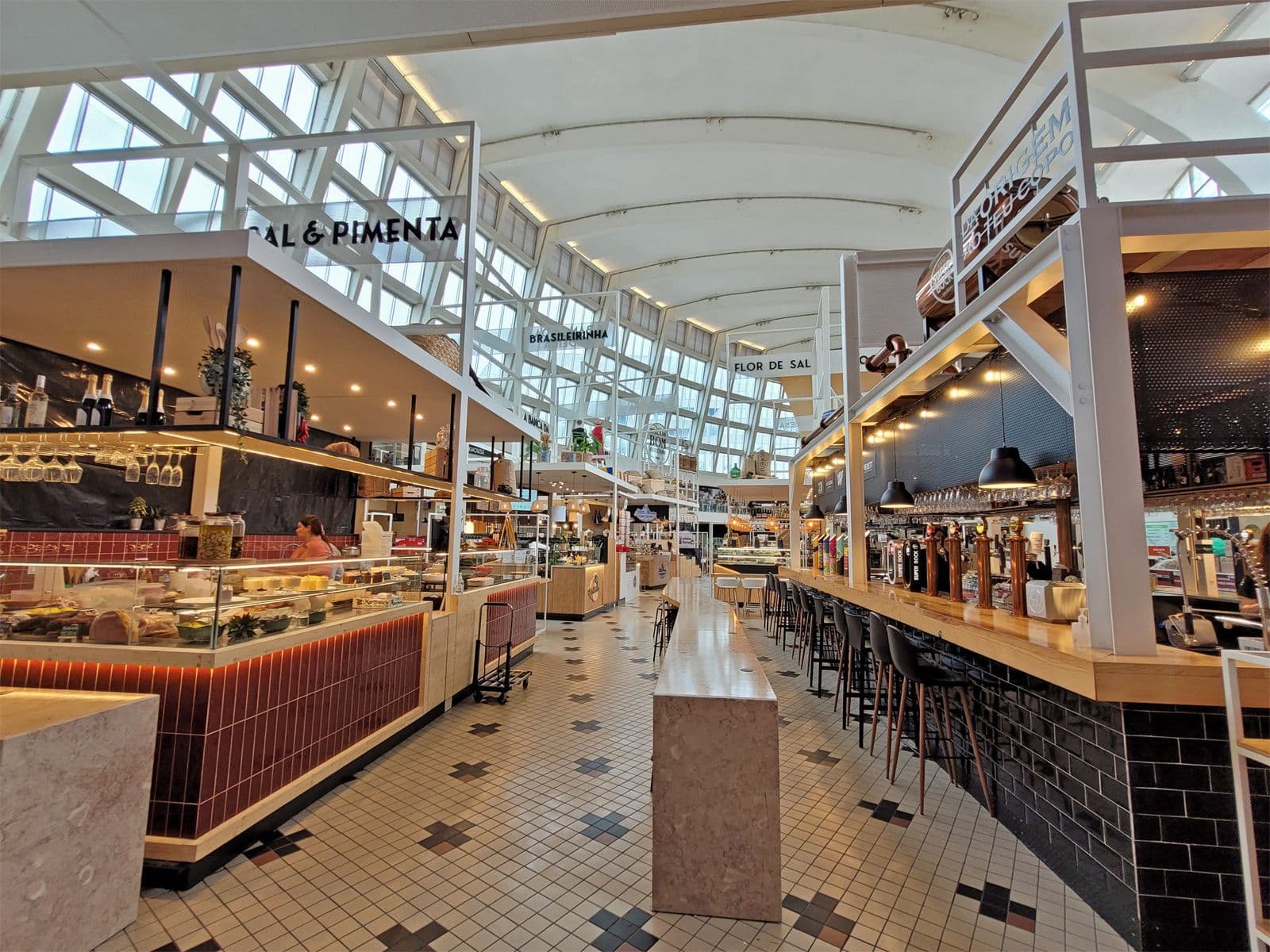 Spacious indoor market with high ceilings, featuring food stalls and a bar with stools. Bright natural light streams through large windows.