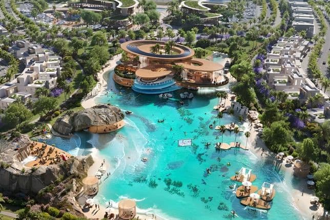 An aerial view of a large lagoon with beaches, rock formations, floating platforms, and a modern clubhouse surrounded by landscaped residential areas.