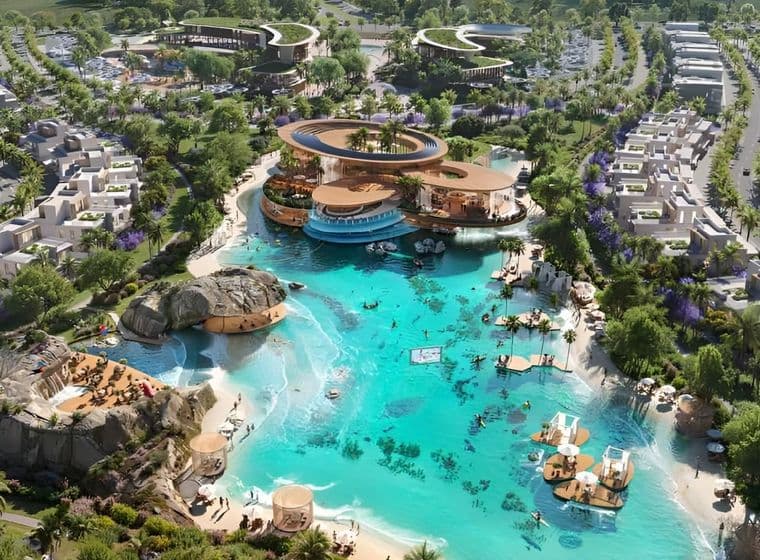 An aerial view of a large lagoon with beaches, rock formations, floating platforms, and a modern clubhouse surrounded by landscaped residential areas.