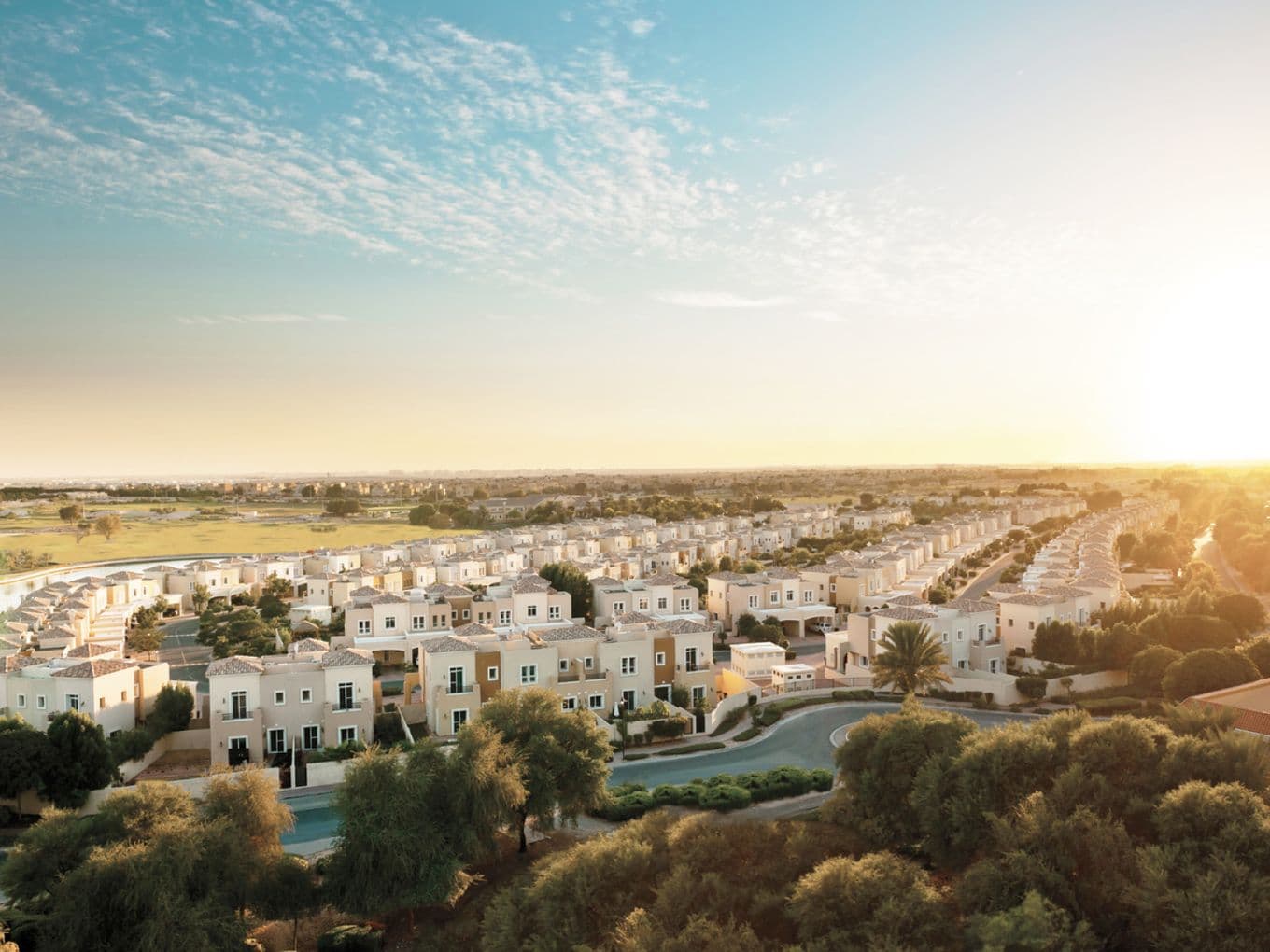 Panoramablick aus der Luft auf die Arabian Ranches Community bei Sonnenuntergang, mit Reihen mediterran gestalteter Villen und üppiger Begrünung, während goldenes Sonnenlicht den Horizont erleuchtet.