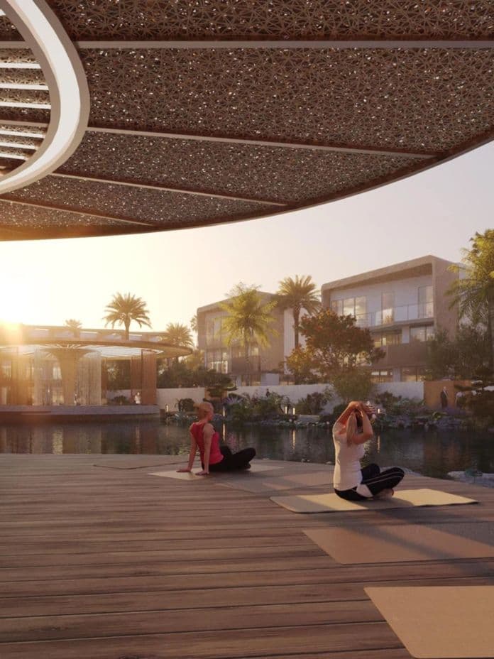 Two people practicing yoga on a wooden deck by a serene lake at sunset, surrounded by palm trees and modern buildings.