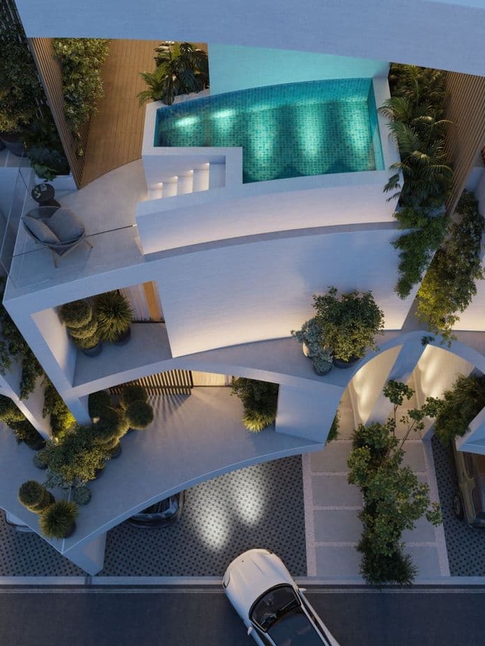 Aerial view of a modern multi-level building with curved balconies, lush greenery, and a rooftop pool, illuminated at night.