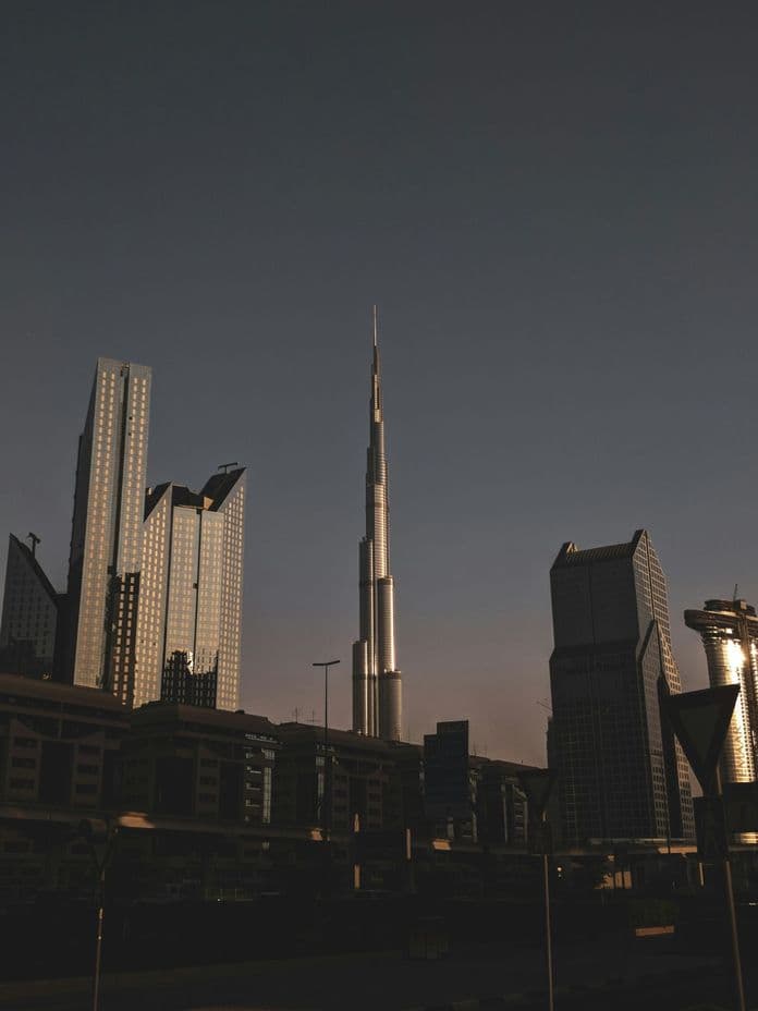 City skyline at dusk with a tall, illuminated skyscraper in the center, surrounded by other modern buildings under a darkening sky.