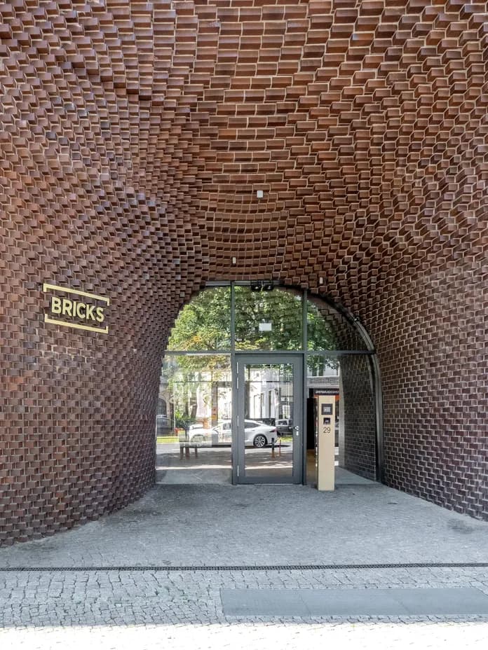 Arched building entrance with honeycomb-like textured brick ceiling, glass double doors reflecting the street, and a yellow BRICKS sign on the left