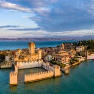 Veduta aerea del Castello Scaligero di Sirmione circondato dalle acque del Lago di Garda, con il borgo storico e la vegetazione rigogliosa sullo sfondo.