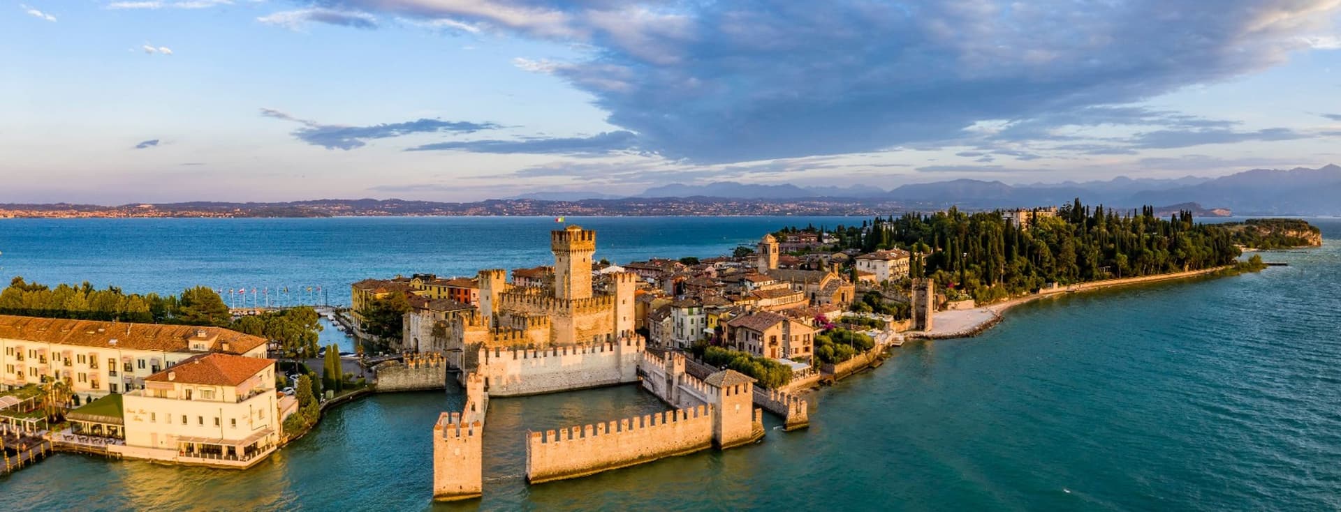 Veduta aerea del Castello Scaligero di Sirmione circondato dalle acque del Lago di Garda, con il borgo storico e la vegetazione rigogliosa sullo sfondo.