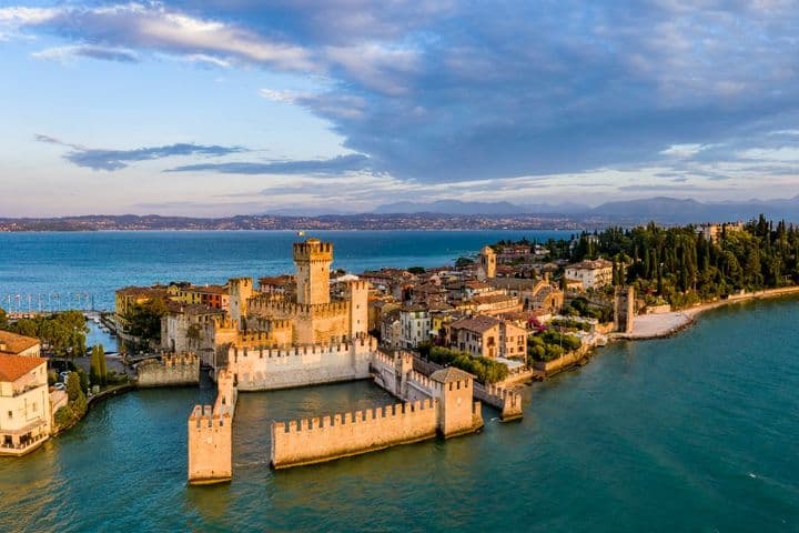 Veduta aerea del Castello Scaligero di Sirmione circondato dalle acque del Lago di Garda, con il borgo storico e la vegetazione rigogliosa sullo sfondo.