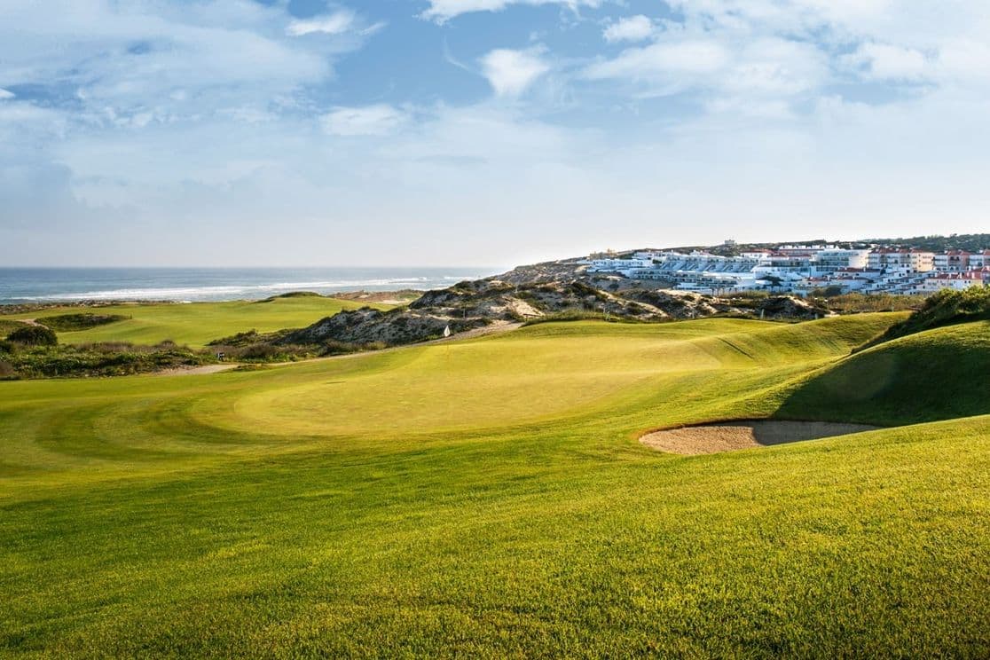 Golf course with lush green fairways, a sand bunker, and ocean view. Houses and hills in the background under a partly cloudy sky.