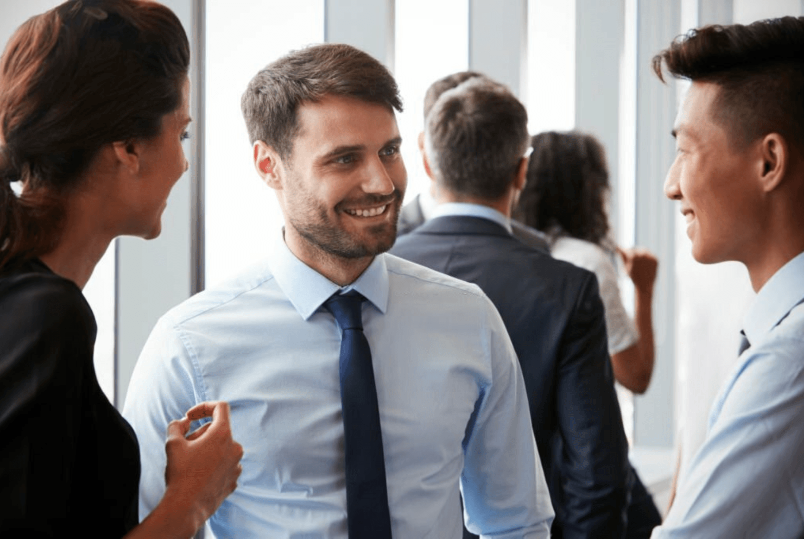 A group of diverse business people in suits are smiling and talking in a bright office setting.