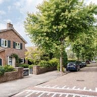 A two-story brick house with dark green shutters sits on a tree-lined street with parked cars.