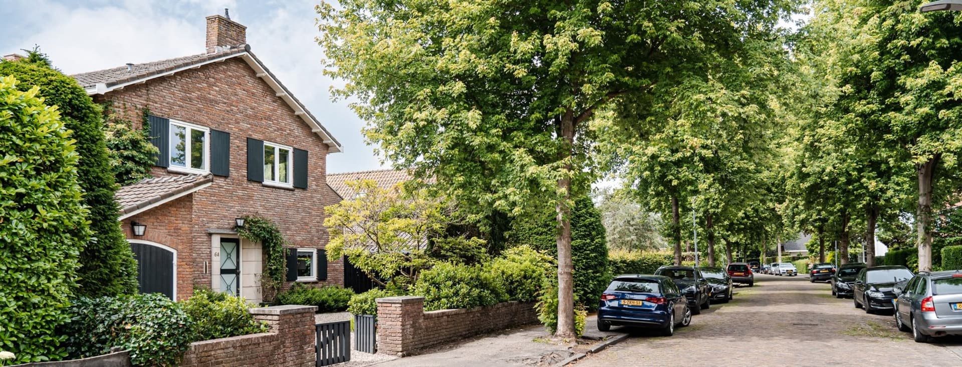 A two-story brick house with dark green shutters sits on a tree-lined street with parked cars.