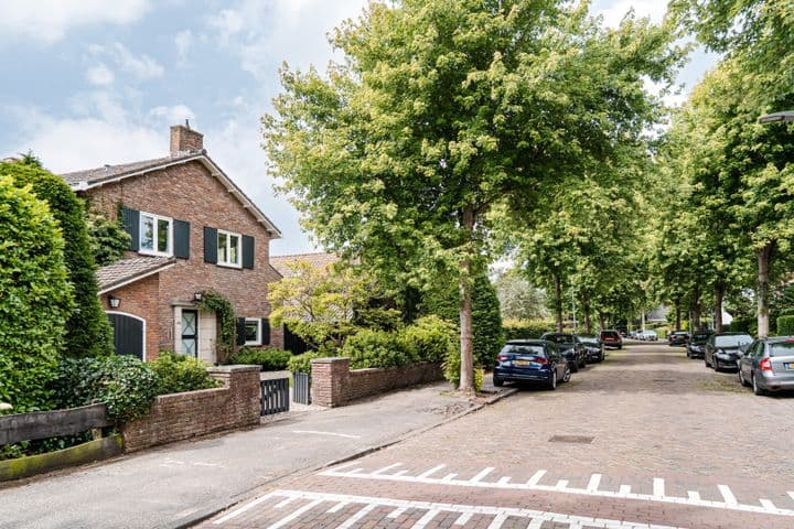 A two-story brick house with dark green shutters sits on a tree-lined street with parked cars.