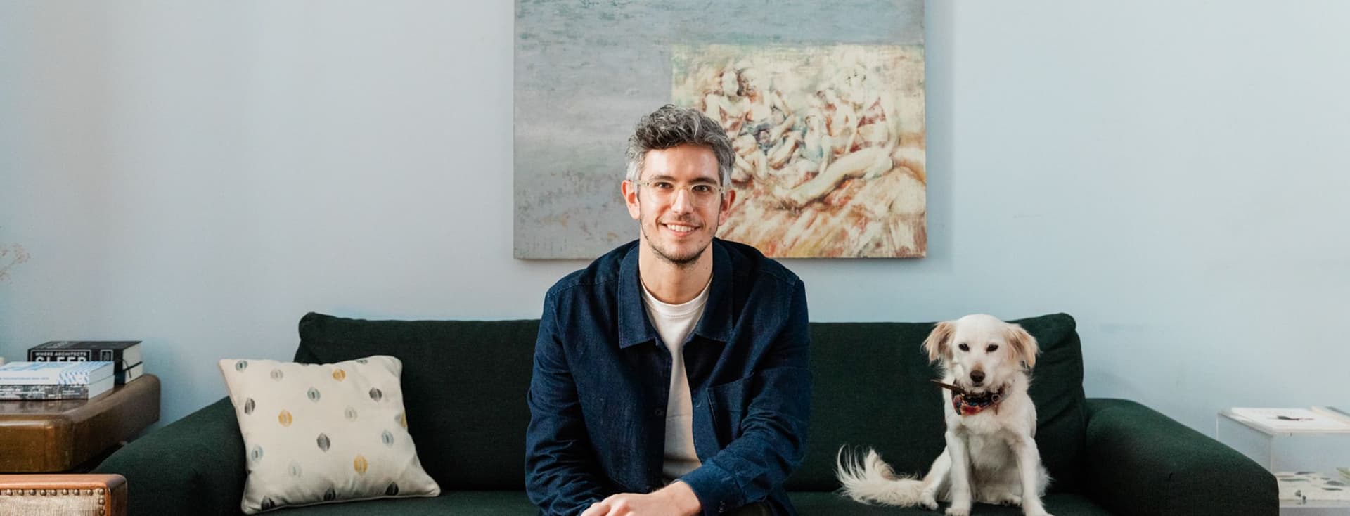 Ένας άνδρας με γυαλιά κάθεται σε έναν πράσινο καναπέ δίπλα σε ένα άσπρο σκύλο σε ένα γαλάζιο δωμάτιο με έναν πίνακα.