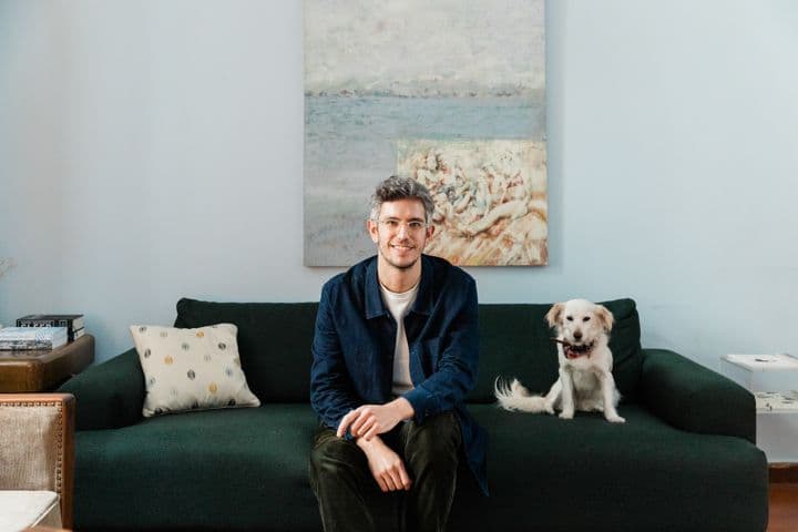 Ένας άνδρας με γυαλιά κάθεται σε έναν πράσινο καναπέ δίπλα σε ένα άσπρο σκύλο σε ένα γαλάζιο δωμάτιο με έναν πίνακα.