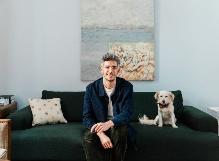 Ένας άνδρας με γυαλιά κάθεται σε έναν πράσινο καναπέ δίπλα σε ένα άσπρο σκύλο σε ένα γαλάζιο δωμάτιο με έναν πίνακα.