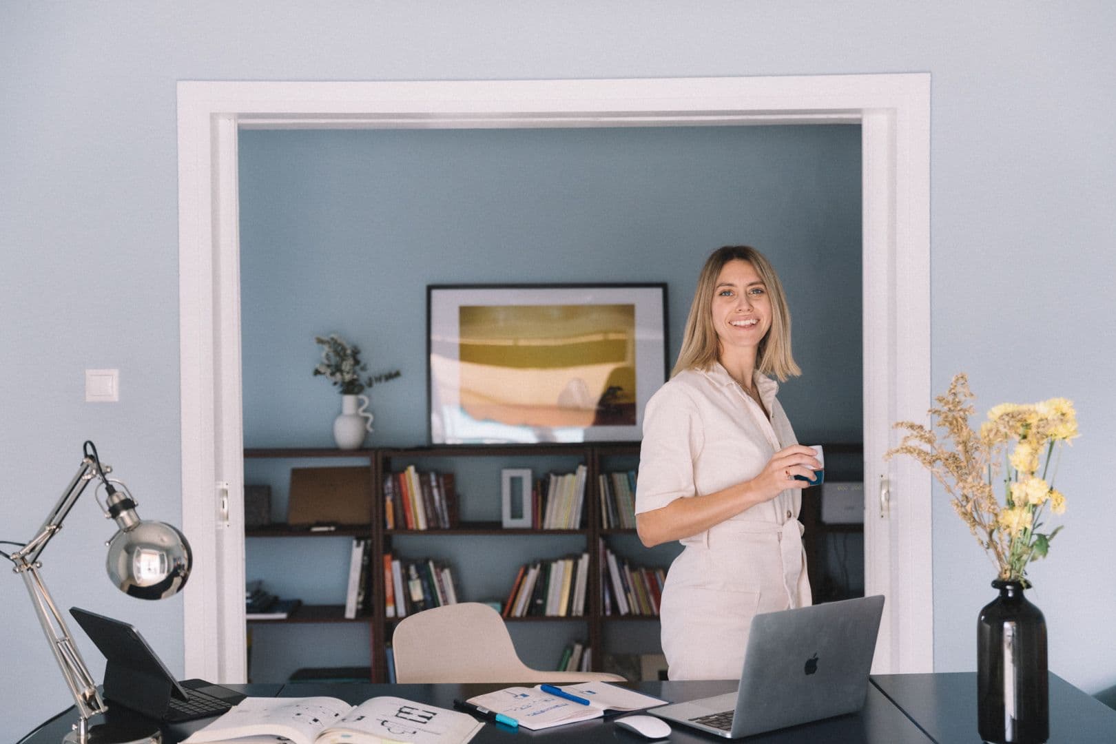 Μια χαμογελαστή γυναίκα με λευκή ολόσωμη φόρμα στέκεται σε μια πόρτα και κρατάει ένα ποτήρι. Ένα γραφείο με φορητούς υπολογιστές και βιβλία βρίσκεται στο προσκήνιο.