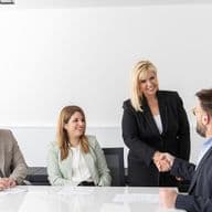 Μια ομάδα πέντε επαγγελματιών κάθονται γύρω από ένα τραπέζι σε ένα φωτεινό γραφείο. Μια όρθια γυναίκα με μαύρο κοστούμι χαμογελάει και σφίγγει τα χέρια με έναν καθισμένο άνδρα, ενώ οι άλλοι παρακολουθούν και χαμογελούν.