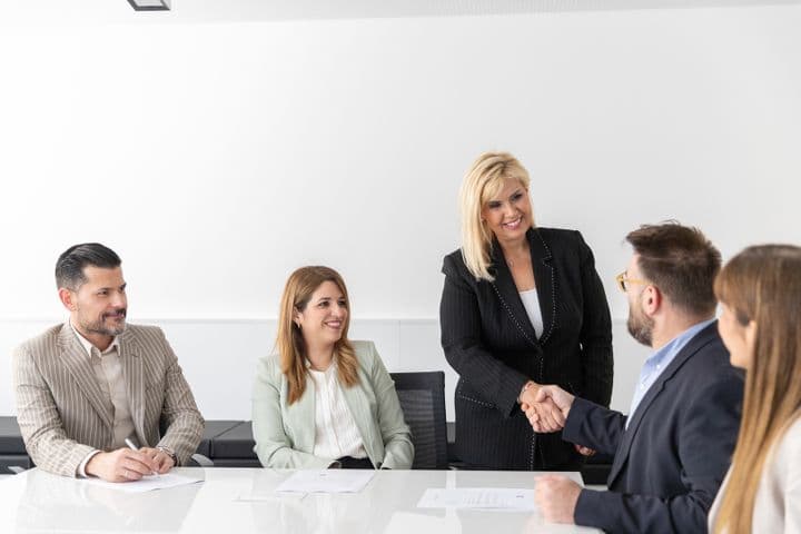 Μια ομάδα πέντε επαγγελματιών κάθονται γύρω από ένα τραπέζι σε ένα φωτεινό γραφείο. Μια όρθια γυναίκα με μαύρο κοστούμι χαμογελάει και σφίγγει τα χέρια με έναν καθισμένο άνδρα, ενώ οι άλλοι παρακολουθούν και χαμογελούν.