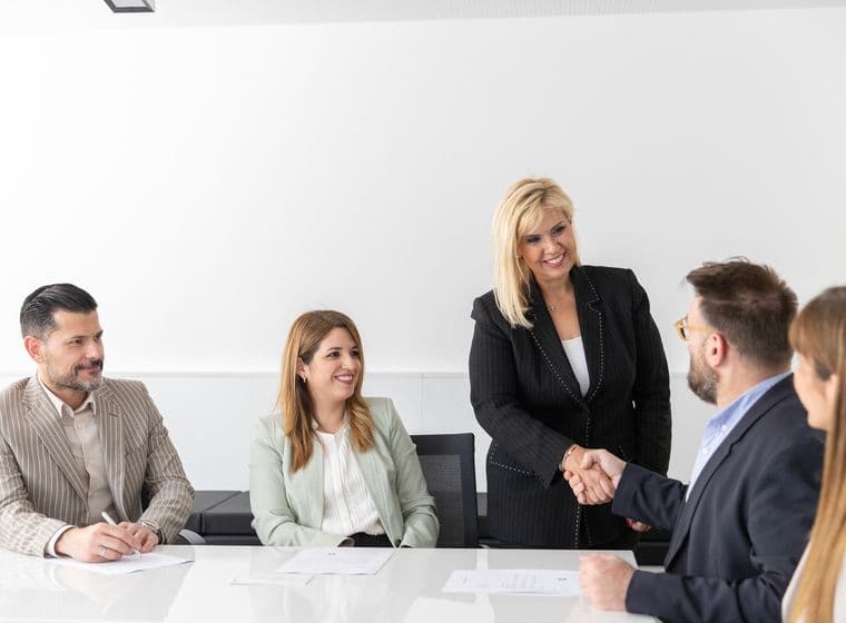Μια ομάδα πέντε επαγγελματιών κάθονται γύρω από ένα τραπέζι σε ένα φωτεινό γραφείο. Μια όρθια γυναίκα με μαύρο κοστούμι χαμογελάει και σφίγγει τα χέρια με έναν καθισμένο άνδρα, ενώ οι άλλοι παρακολουθούν και χαμογελούν.