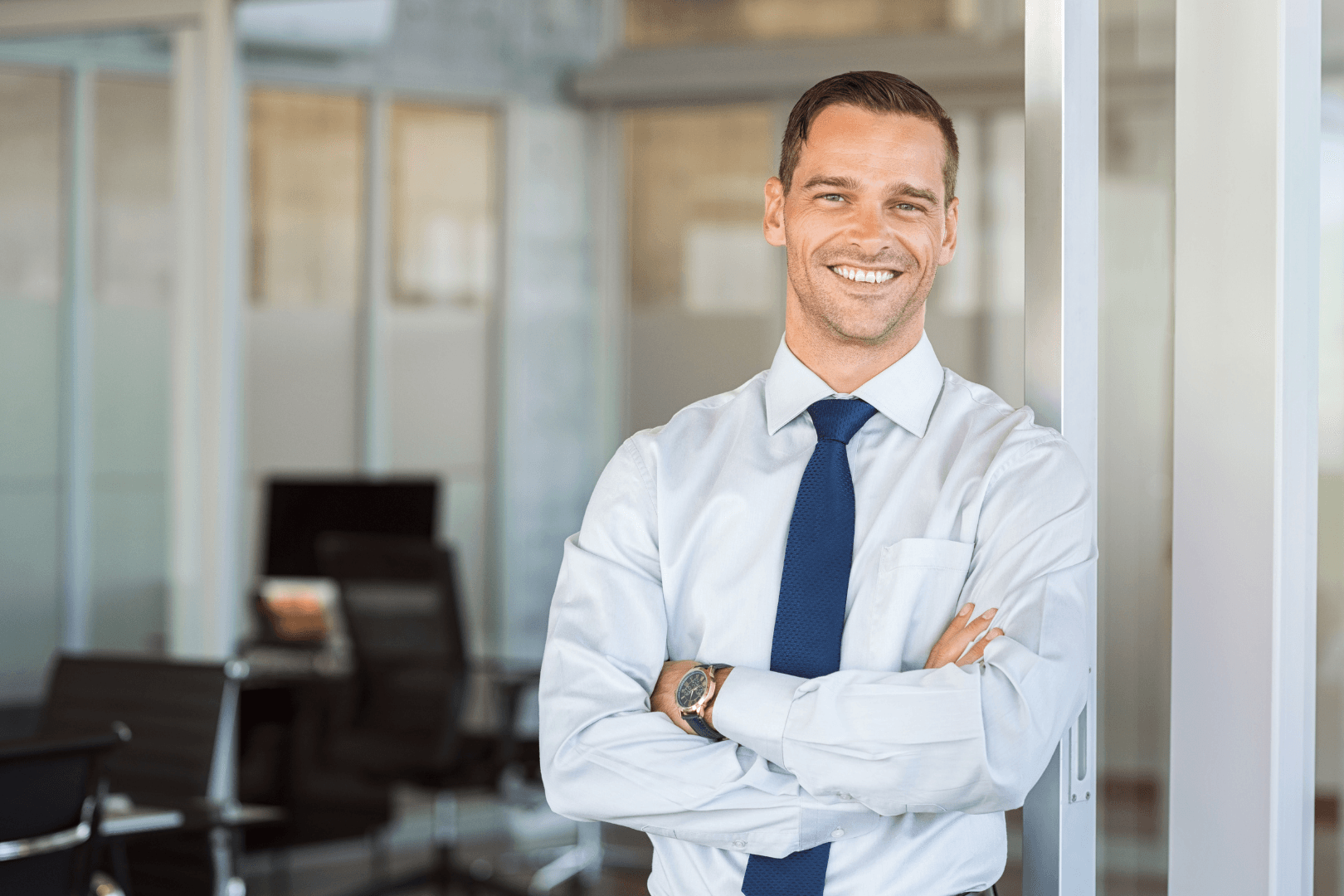A smiling man in a white shirt and blue tie stands with arms crossed in a modern office.