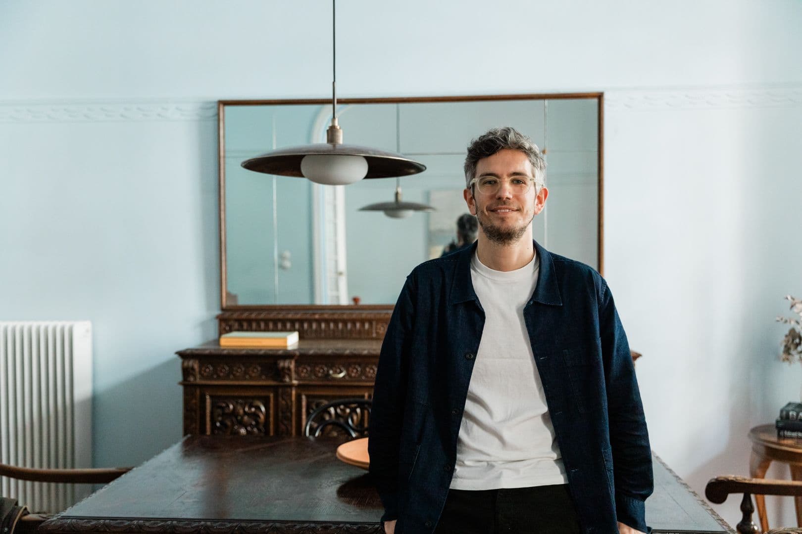 Ένας χαμογελαστός άντρας με γυαλιά και ένα μπλε μπουφάν στέκεται σε ένα γαλάζιο δωμάτιο με αντίκες έπιπλα και έναν μεγάλο καθρέφτη.