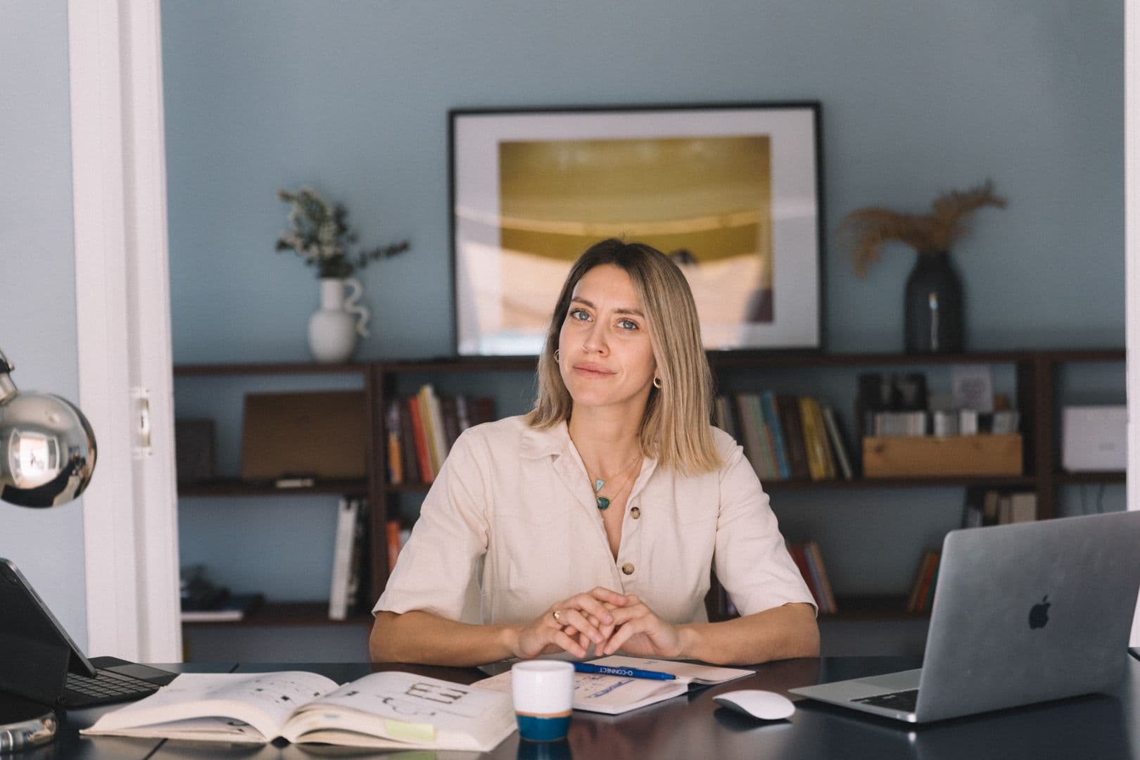 Γυναίκα με ξανθά μαλλιά κάθεται σε ένα γραφείο με ένα λάπτοπ, ένα βιβλίο και μια κούπα καφέ. Βιβλιοθήκες και έργα τέχνης βρίσκονται στο φόντο.