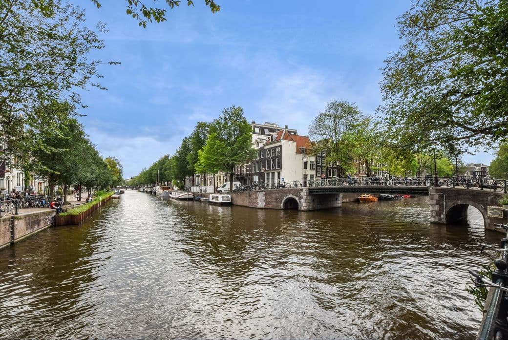 Amsterdam canal view with brick bridges, boats, and bikes. Buildings line the canal under a blue sky with trees.