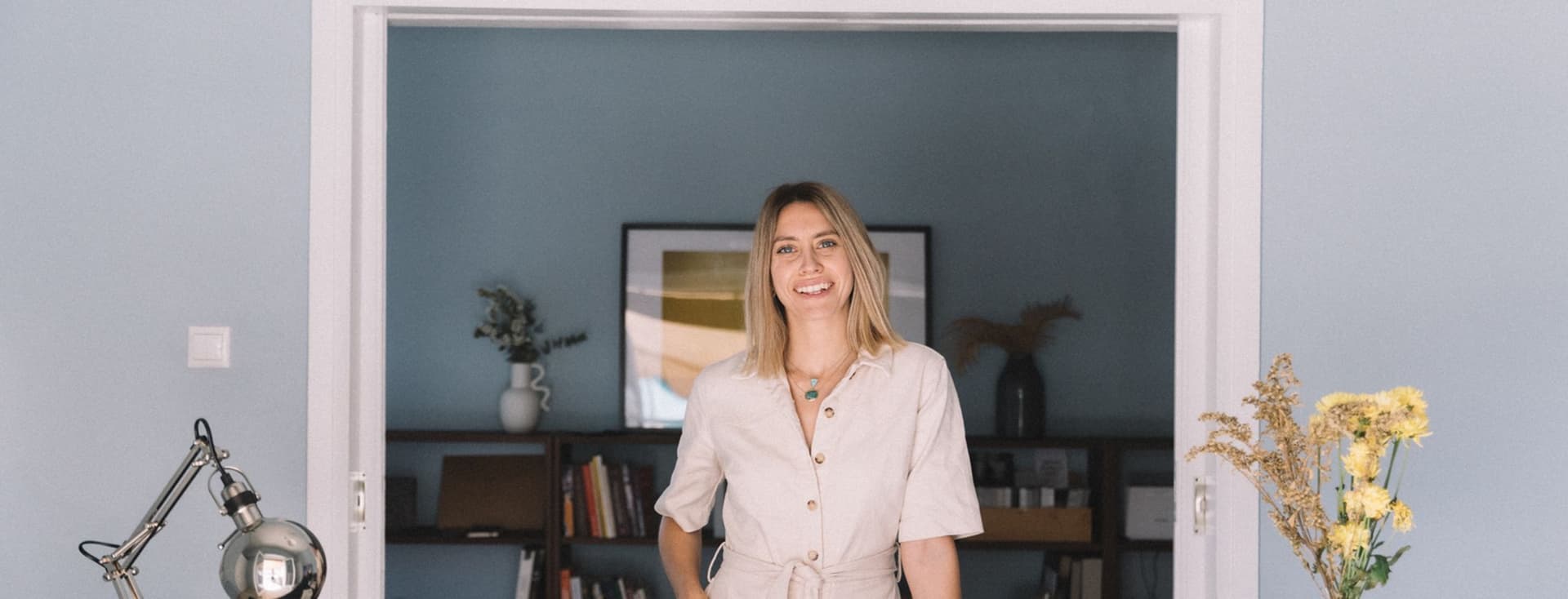 Μια χαμογελαστή γυναίκα με μπεζ φόρμα στέκεται σε ένα γραφείο με ένα λάπτοπ και λουλούδια σε ένα μπλε δωμάτιο.