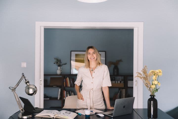Μια χαμογελαστή γυναίκα με μπεζ φόρμα στέκεται σε ένα γραφείο με ένα λάπτοπ και λουλούδια σε ένα μπλε δωμάτιο.